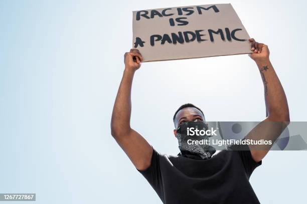 Low Angle View Of African American Man With Scarf On Face Holding Placard With Racism Is A Pandemic Lettering Against Blue Sky Stock Photo - Download Image Now