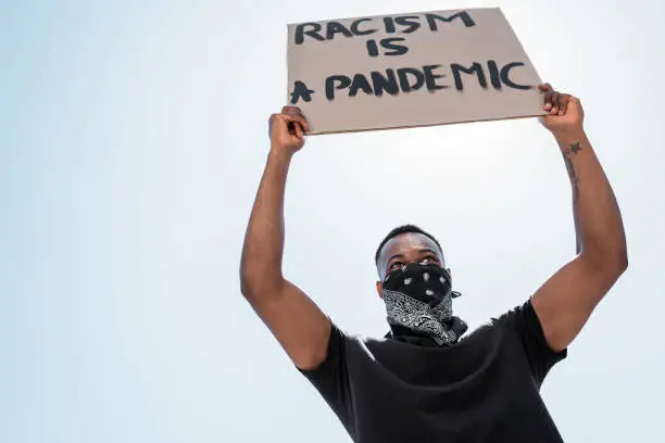 Photo of low angle view of african american man with scarf on face holding placard with racism is a pandemic lettering against blue sky