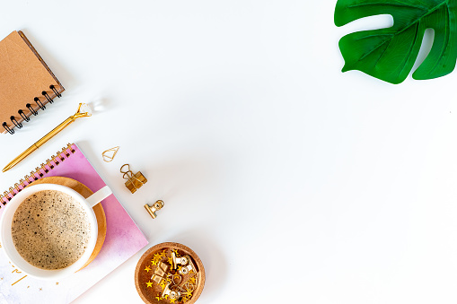 Flat lay of white working table place background with cup of coffee putting on it. Top view glasses, leaf, golden paper binder clips, Notebook and pen. Desktop mock up planner with copy space.