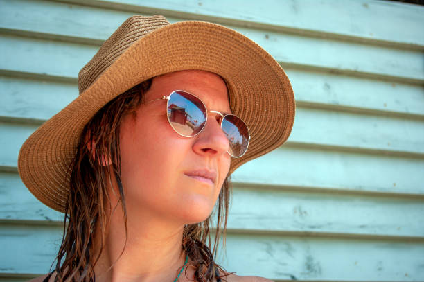 Young woman in a straw hat with sunglasses and looks forward \nwith glasses reflecting a small beach house