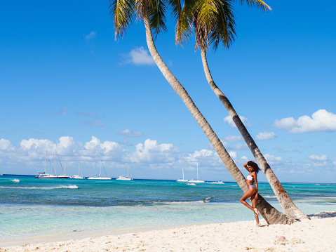 Landscape of an exotic, well-preserved and clean beach where an athlethic woman poses seductively by plam trees.