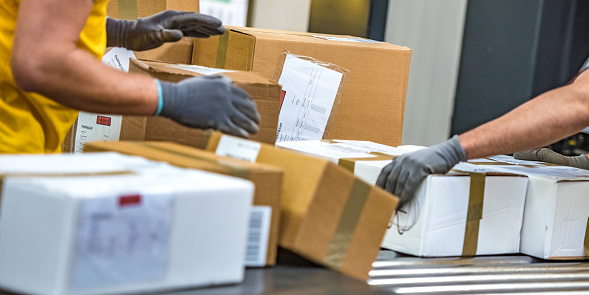 Close-up photo of postal office worker's hands handling packages at a conveyor belt in a warehouse.