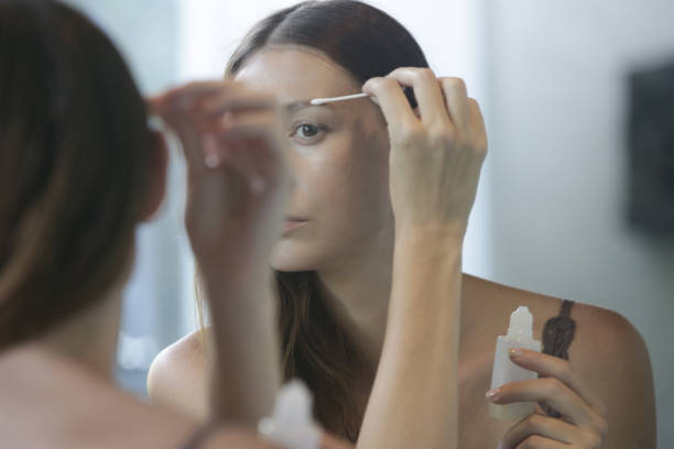 jeune femme caucasienne appliquant le sérum pour la croissance de sourcils et de cils devant le miroir. routine de beauté de nuit. - prélèvement de sérum photos et images de collection