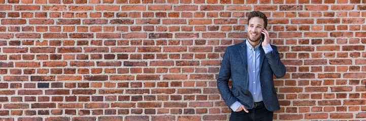 Business man talking on mobile phone panoramic banner of brick wall background texture. Happy young businessman using cellphone in urban setting wearing smart casual suit.
