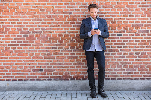 Businessman sms texting phone app in city street on brick wall background. Business man holding smartphone in smart casual wear standing. Urban young professional lifestyle.