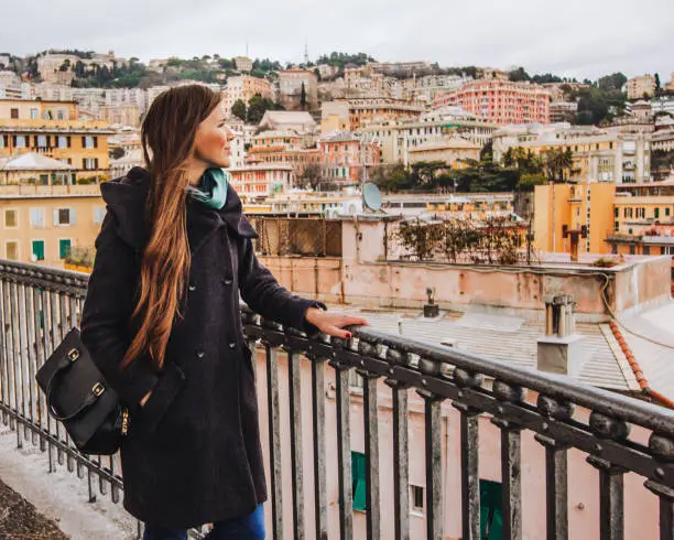 A girl with long hair in a coat looks at the panorama of Genoa, cute houses and roofs