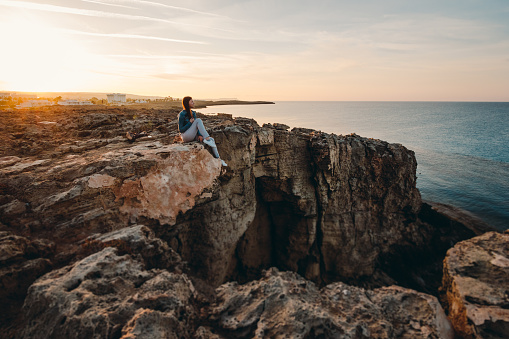 Woman traveler sitting near sea on top of cliff in summer mountains and enjoying view of nature and sea. Cape Greco, Cyprus, Mediterranean Sea. Sunrise.