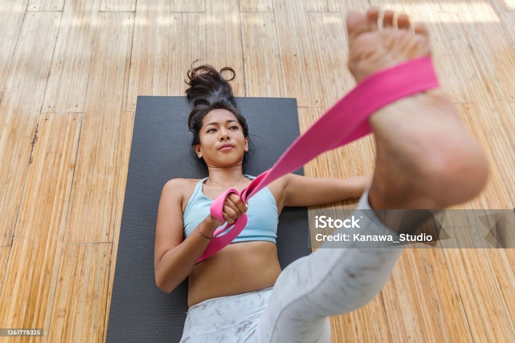 Young Asian woman using yoga strap to exercise flexibility Shot of a beautiful young Asian woman lying down on the floor, using a pink yoga strap to practice flexibility at her home Strap Stock Photo