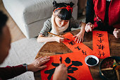 Grandparents practising Chinese calligraphy for Chinese New Year Fai Chun (Auspicious Messages) and teaching their granddaughter by writing it on couplets at home