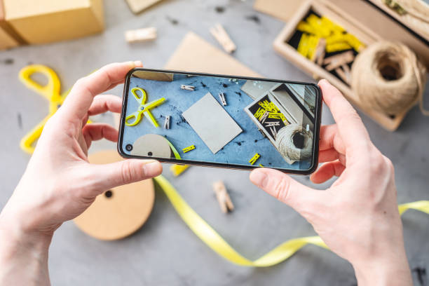 istock Women's hands take photos on a mobile phone. There are tools for creating handmade gift packaging on the gray background. Craft paper, bright yellow ribbon and other things for creativity. 1267808328