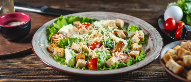 istock Plate with appetizing fresh Caesar salad on a wooden table and ingredients for cooking it. Top view 1267847587