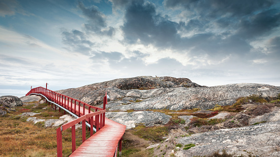 Red wooden walkway along the natural rocky Ilulissat Icefjord Coast under dramatic twilight skyscape, Panorama view along the greenlandic artic coast covered with green mosses. Ilulissat Icefjord Coast, Ilulissat, Greenland, Denmark, Arctic Polar Region