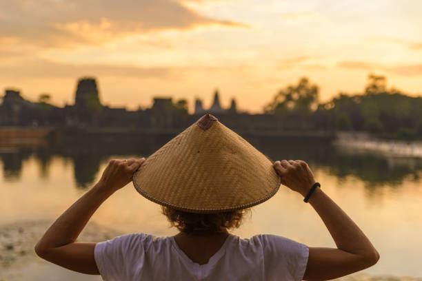 ein tourist besucht angkor wat ruinen bei sonnenaufgang, reiseziel kambodscha. reflexion auf wasserteich inmitten des dschungels, frau mit traditionellem hut, rückansicht. - cambodia traditional culture ancient angkor stock-fotos und bilder