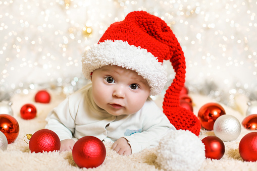 Adorable baby in a red Christmas hat around red balls on a white background