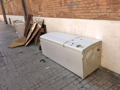 Valencia, Spain - August 21, 2020: Abandoned refrigerator lying on the sidewalk. Usually people don't know where to leave household appliances so they leave them in the street next to the garbage bins hoping that the collectors will grab them