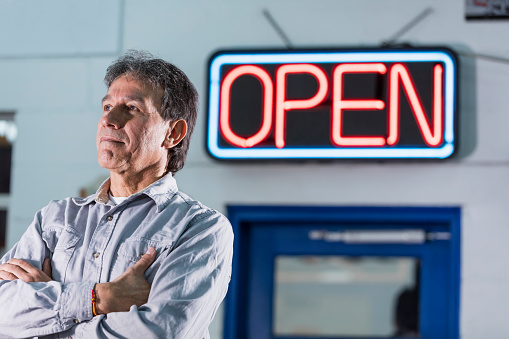 A senior Hispanic man in his 60s, a small business owner, standing with his arms crossed in front of a large, neon, open sign. His repair shop is open for business, and he has a serious but hopeful expression on his face.