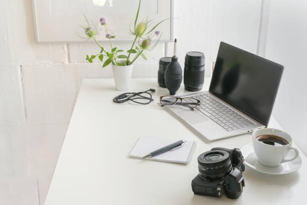 istock White office desk with laptop, camera, lenses, cable, glasses, notepad and a coffee cup, modern workplace of a photographer against a bright wall, copy space, selected focus 1267949625