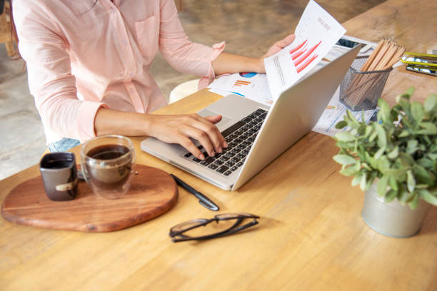 Woman working at home office desk using laptop business financial document chart and graph on wooden table with coffee cup. Woman freelance reading business graphs sale report on business office desk mujer que trabaja en el escritorio de la oficina en casa utilizando la tabla de documentos financieros de negocios portátiles y el gráfico sobre la mesa de madera con taza de café. mujer freelance leyendo gráficos de negocios informe de venta en oficin