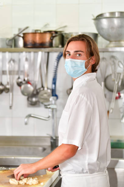 Young male chef with a surgical mask using a knife to cut potatoes on a cutting board while looking at the camera