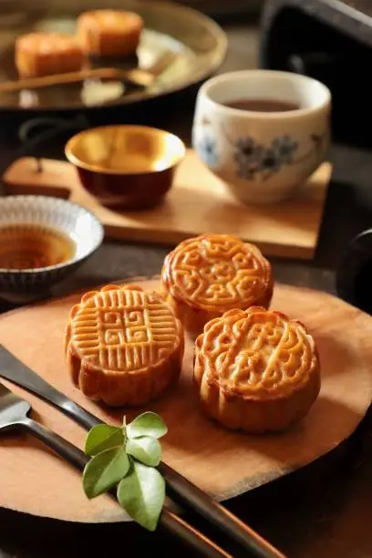 Mooncake, the traditional Chinese pastry made seasonally for the Chinese Mid-Autumn Festival. The cakes are filled with lotus seed paste or red bean paste; then moulded with various ornamental patterns. One pattern has some Chinese characters in it; which means Mid-Autumn. The mooncakes are plated on a small cutting block. A set of fork and knife is placed on the plate. Sprigs of orange jasmine leaves are used as garnish.