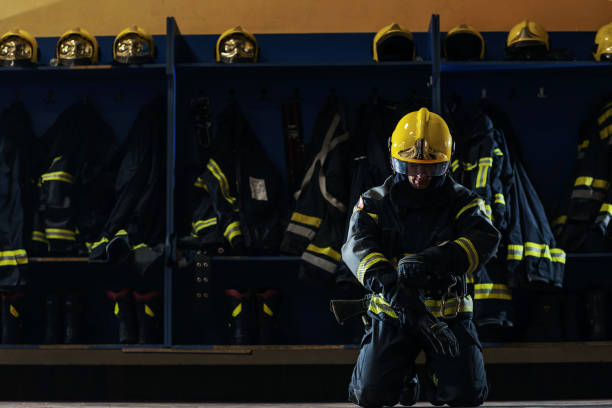 istock Brave young firefighter in protective uniform kneeling and putting on gloves on head and preparing for action. Fire station interior. 1268017742
