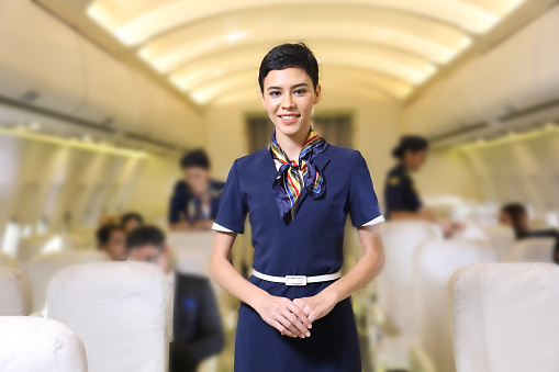 Caucasian flight attendant posing with smile at middle of the aisle inside aircraft with passengers seated on the background