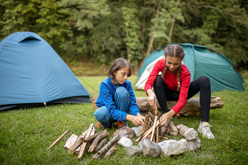 Boy and girl having a fun on camping in nature near forest and preparing campfire in front of their tents.