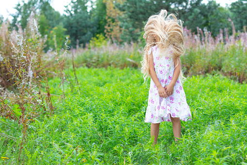 A girl in a white sundress, with lush blond hair, stands in a flowering meadow, shook her head, no face is visible, her hands are folded. Lush mane of hair. Childhood, fun, reunion with nature.