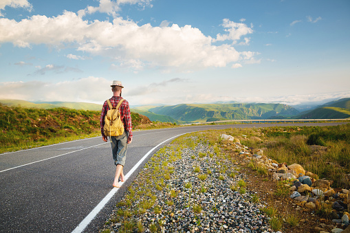 A young bearded hipster in a shirt hat and barefoot walks along a country road in the mountains. Hitchhiking and free travel.