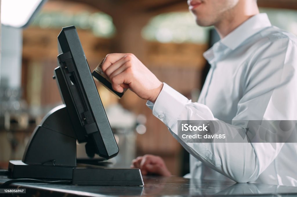 small business, people and service concept - happy man or waiter in apron at counter with cashbox working at bar or coffee shop. small business, people and service concept - happy man or waiter in apron at counter with cashbox working at bar or coffee shop Point Of Sale Stock Photo
