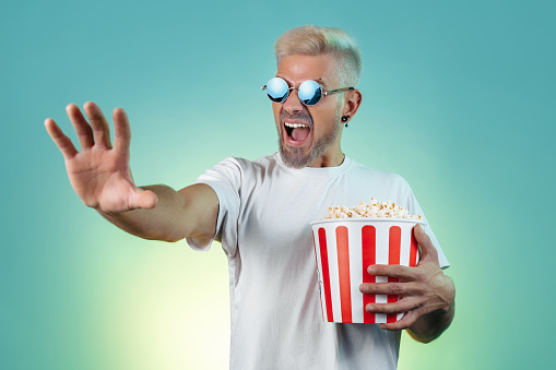 Excited hipster blonde man wearing stylish sunglasses and holding bucket with popcorn in studio in neon lights