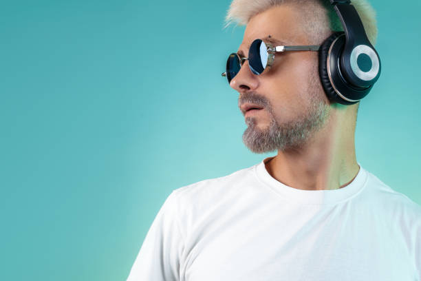 シアンの背景にスタイリッシュなヘッドフォンで音楽を聴くサングラスを身に着けているスタイリッシュなひげを生やしたヒップスターの若者。 - free standing audio ストックフォトと画像