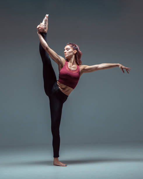 istock Flexible young female dancer practices stretching in stylish sportswear barefoot in studio isolated on gray background 1268061704