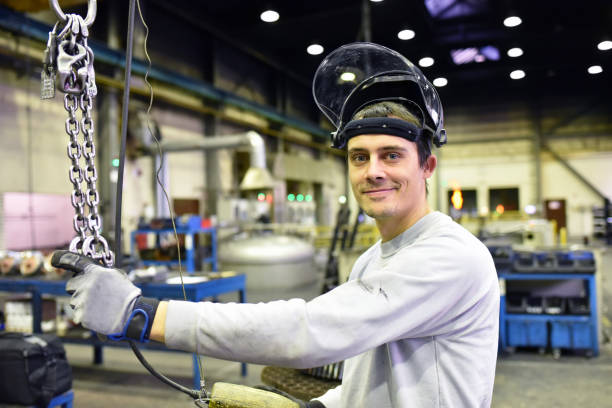 彼の職場の近代的な工業ホールでヘルメットを持つ若い笑顔の産業労働者の肖像画 - 金属工 ストックフォトと画像