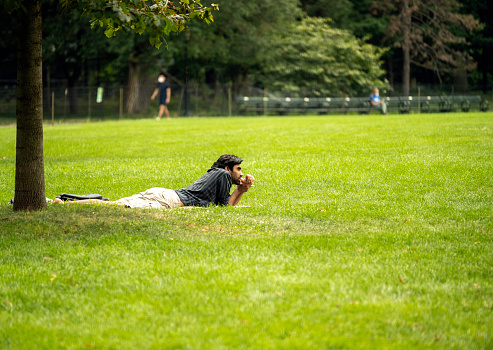 New York, NY, USA - August 23, 2020: A man holds a pen and lies in the grass as he looks out over the Great Lawn in Manhattan's Central Park on a hot summer day.