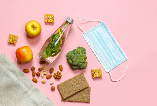 Layout with a protective mask, a bottle of water, apples, broccoli and nuts laid out of the bag. pink background with space stock photo