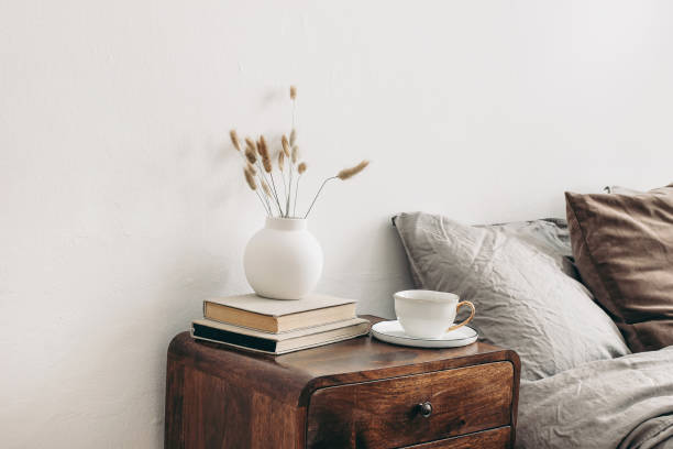 Modern white ceramic vase with dry Lagurus ovatus grass and cup of coffee on retro wooden bedside table. Beige linen and velvet pillows in bedroom. Scandinavian interior. Homestaging. Modern white ceramic vase with dry Lagurus ovatus grass and cup of coffee on retro wooden bedside table. Beige linen and velvet pillows in bedroom, scandinavian interior. Homestaging. mahogany photos stock pictures, royalty-free photos & images