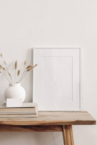 istock Portrait white picture frame mockup on vintage bench, table. Modern ceramic vase with dry Lagurus ovatus grass, books and busines card. White wall background. Scandinavian interior. Vertical. 1268077664