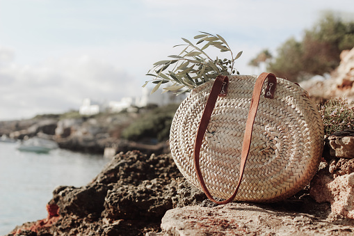Summer vacation still life. French straw basket with olive tree branches on beach rocks. Blurred background with sea, white houses and boat. Selective focus. Mediterranean Mallorca island travel.