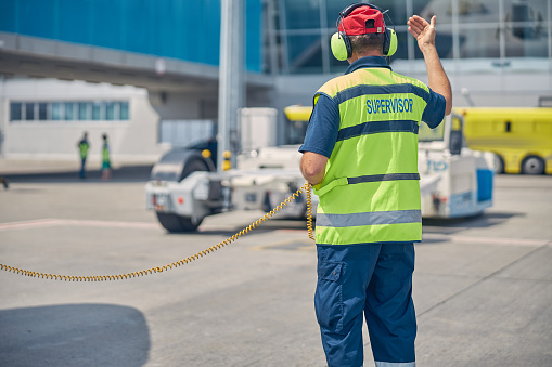 Back view of a man in a safety vest and headphones directing a vehicle operator