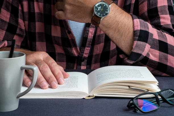 A man in a plaid shirt is reading a book at the table. Glasses, watch and cup. A man in a plaid shirt is reading a book at the table. Glasses, watch and cup. coffee table top stock pictures, royalty-free photos & images