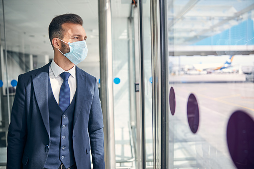 Handsome elegant man in a suit wearing a medical mask and looking through the glass door of an airport. Website banner