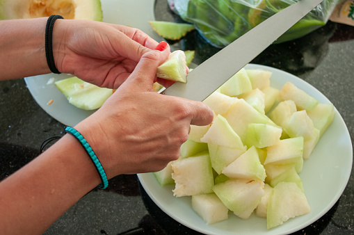 Young Woman hands cutting honeydew melon with large kitchen knife on black kitchen countertop