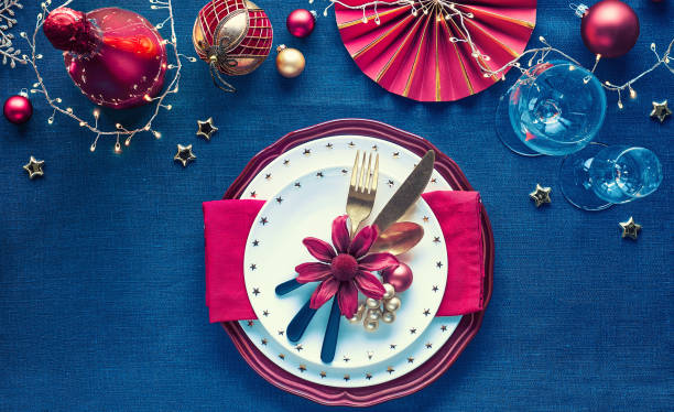 istock Christmas table setup with golden, pink and red decor on classic blue linen textile. Plates with flowers, utensils and vine glass. 1268116671