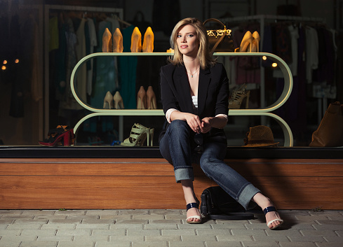 Young fashion blond business woman sitting on the mall window Stylish female model in black blazer