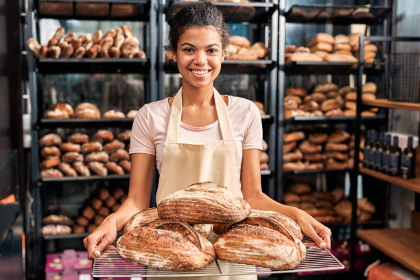 petite entreprise. jeune femme dans le tablier au magasin de boulangerie retenant des pains posant à l’appareil-photo heureux - premier emploi photos et images de collection