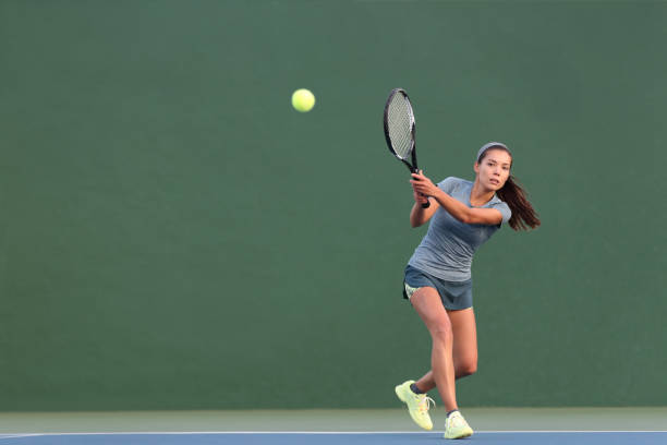 tennis playing woman hitting ball on green hard court. asian athlete girl returning serve with racket wearing skort and shoes - womens tennis imagens e fotografias de stock
