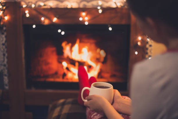 Cup of tea in woman's hands sitting near fireplace Close up cup of hot tea in woman's hands sitting near fireplace with festive Christmas lights in cozy room. holidays and seasonal stock pictures, royalty-free photos & images