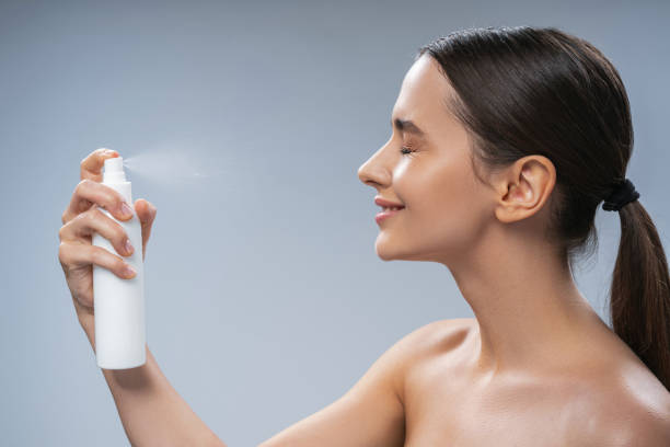 Smiling female with a ponytail doing a beauty procedure Side view of a pleased young dark-haired Caucasian woman spraying thermal water on her face spraying water stock pictures, royalty-free photos & images