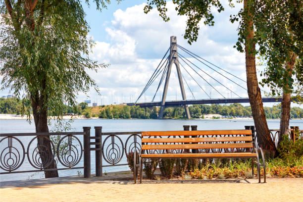 Summer landscape with a wooden bench on the Dnipro embankment against the background of the Northern bridge over the river in blur. Park wooden bench on the Dnipro embankment against the background of a summer landscape and the Northern bridge over the river in Kyiv. dnipropetrovsk stock pictures, royalty-free photos & images